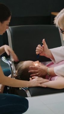 Dikey video: Sağlık merkezinin lobisinde el feneriyle küçük baygın bir kıza kıdemli sıhhiye danışmanı. Hastalıklı küçük bir çocuğa yardım etmeye çalışan, hastanedeki sandalyelerde bayılan doktor.