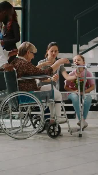 垂直录像 坐在轮椅上的老年妇女在候车室与小女孩交谈 家人来看老年残疾患者 轮椅使用者在医院试图用步行架走路 — 图库视频影像