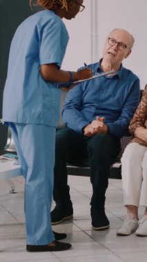 Dikey video: Afrikalı Amerikalı hemşire bekleme odasında yaşlılarla konuşuyor, doktorla tıbbi konsültasyona katılmak için bekliyor. Muayeneden önce kontrol raporunu doldurma asistanı