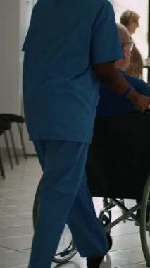 Dikey video: Tekerlekli sandalyedeki hastaya klinik lobisinde ailesiyle buluşması için yardım eden hemşire, klinikte kronik bozuklukla uğraşan eski bir hasta. Anne ve çocuk bekleme odasını ziyaret ediyor..