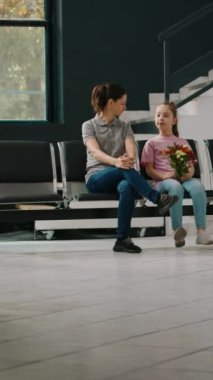 Dikey video: Anne ve çocuk tıbbi tesiste tekerlekli sandalyedeki yaşlı kadını ziyaret ediyor, engelli kıdemli bir hastayla konuşuyor. Hastane resepsiyonunda büyükanneme çiçek getiren küçük kız.