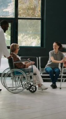 Dikey video: Tekerlekli sandalyede oturan yaşlı bir yetişkin bekleme salonunda küçük bir çocukla konuşuyor, ailesi yaşlı bir hastayı ziyarete geliyor. Tekerlekli sandalye kullanıcısı hastanede yürümeye çalışıyor.