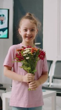 Dikey video: Hastane bekleme salonunda çiçek buketi tutan gülümseyen küçük kız, sağlık merkezindeki eski hastayı ziyaret etmeye hazırlanıyor. Küçük çocuk anne ve babasıyla bekleme odasında oturuyor..