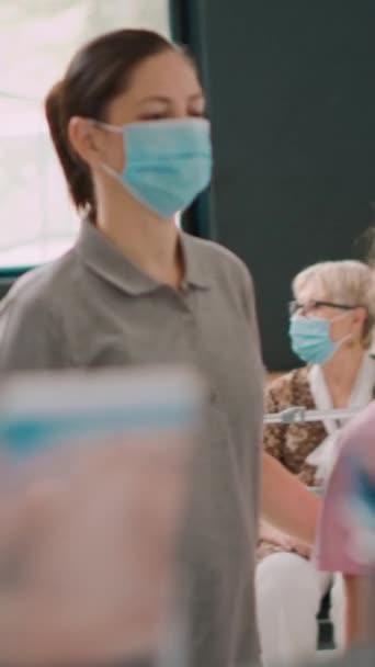 垂直ビデオ 病院のカウンターで受付係と話す顔のマスクを持つ母親と子供 一般開業医を見るために医療の約束を持っている人々 受付で待っている女と女 — ストック動画