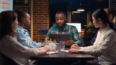 Afrika kökenli Amerikalı analitik uzmanı iş toplantısında laptop ekranında veri görselleştirmesi gösteriyor. Takım çalışması, çeşitli iş arkadaşları şirket raporlarını gece vakti tartışıyor.