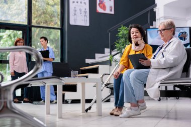 Rahim boynu tasması olan uzman bir hasta not almak için tablet kullanıyor, hastane bekleme odasında check-up yapıyor. Fiziksel ağrısı ve yarası olan Asyalı bir kadın randevusu var..