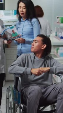 Dikey video: Tekerlekli sandalyedeki erkek müşteri ve eczacı eczaneden ilaç almak için eczacıyla konuşuyor. Kronik engelli kişi tıbbi yardım ve vitamin alıyor, satın alıyor