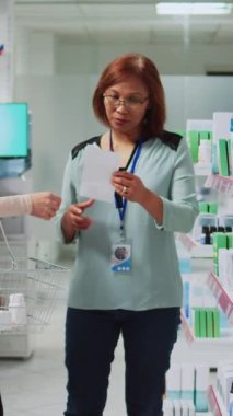 Dikey video: Genç eczacı müşteriye ilaç ve vitamin vermek için eczanede reçete kağıdı okuyor. Eczacıdan takviye, hap ya da ilaç kutuları alan Asyalı kişi.