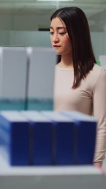 Dikey video: Asyalı müşteri eczane raflarındaki ilaç kutularını kontrol ediyor, tedavi ve hap bulmak için paketlere bakıyor. Tıbbi malzemeler ve ilaç ürünleri satın almak için broşür okumak
