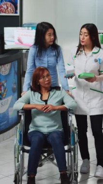 Dikey video: Tekerlekli sandalyedeki kadın hasta bakıcı ve kadın eczanede reçeteli ilaç ya da haplar hakkında uzman soruyorlar. Sosyal hizmet görevlisi engelli kişilere yardım