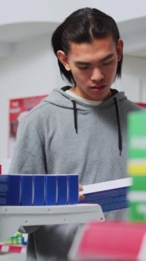 Dikey video: Asyalı kişi eczanede tıbbi ürünlerin kutularını işaretliyor, raflardan ilaç ve vitamin alıyor. Erkek müşteri ilaç paketleri üzerinde broşür okuyor, ilaç arıyor.