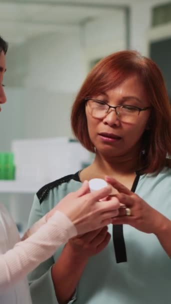 垂直ビデオ 薬局でアジアの女性に循環器系薬を示す医療コンサルタント 心臓の問題を治療するための治療を説明 医療製品についてクライアントと話す女性薬剤師 — ストック動画