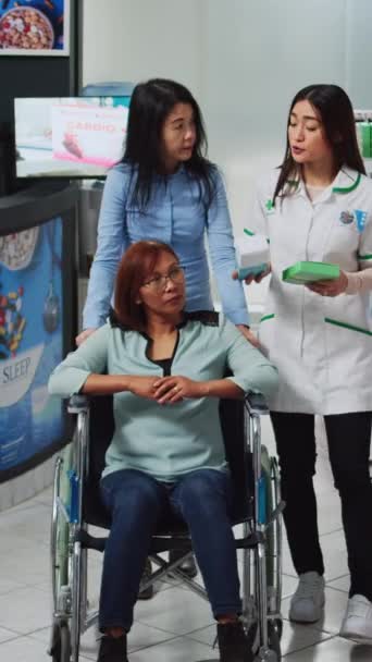 垂直ビデオ 車椅子の女性管理者と女性が薬や薬について専門家に尋ねる 薬局でプレクリピオン薬を探しています 障害者を支援するソーシャルワーカー — ストック動画