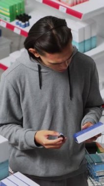 Dikey video: Genç bir adam eczane dükkanında kutu kutu uyuşturucuya bakıyor, reçeteli ilaç almak için paket okuyor. Raflardan tedavi edilmeden önce ilaç ve takviyeleri kontrol ediyorum. El bilgisayarı