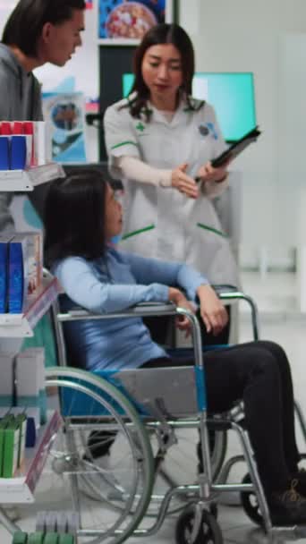 垂直ビデオ 薬局の従業員は車椅子の女性に薬箱を与えます 障害のある人を助ける男性管理者 薬局の顧客やソーシャルワーカーに薬を与える薬剤師 — ストック動画