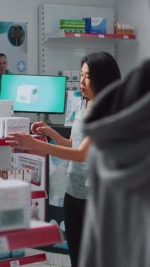 Dikey video: Eczanede ilaç arayan kadın müşteri, ilaç kutuları ve şişelerle dolu raflara bakıyor. İlaç paketleri, ilaç ürünleri okuyan eczane müşterisi