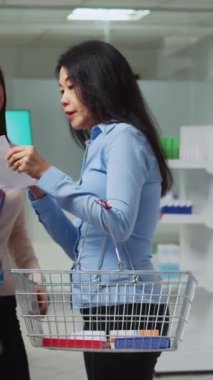 Dikey video: Eczane çalışanı müşteriye ilaç ve hap vermek için eczanede reçete kağıdı okuyor. Eczacıdan takviye, ilaç ya da ilaç kutuları alan Asyalı kişi.