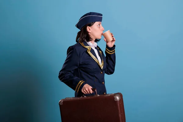 Stewardess in professional airline uniform carrying baggage, while drinking coffee to go. Flight attendant with luggage in airport, air hostess with suitcase holding tea paper cup, side view