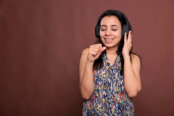 Smiling indian woman in headphones, singing in imaginable microphone, listening to music with satisfied facial expression. Lady with closed eyes holding mic, wearing earphones, studio mid shot