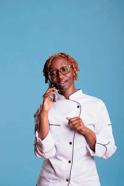 身着厨师制服的非洲裔美国女人和工作室里的同事们进行着愉快的交谈 专业女厨师使用蓝色背景移动设备 — 图库照片