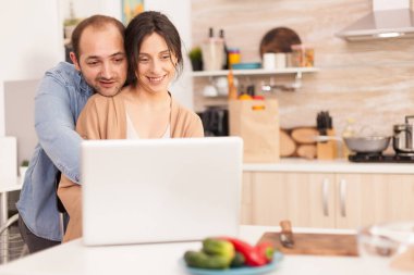 Erkek arkadaşı dizüstü bilgisayar kullanırken kız arkadaşına mutfakta sarılıyor. Mutlu ve neşeli bir romantik çift evlerinde modern kablosuz internet teknolojisini kullanıyor.