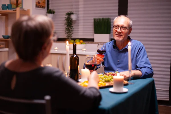 Senior man telling a story to his wife while celebrating in kitchen with wine and food. Senior couple sitting at the table in dining room , talking, enjoying the meal, celebrating their anniversary in