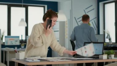 Busy woman in startup office talking on smartphone looking at papers with sales charts at desk. Business analyst using mobile phone searching for data statistics with laptop showing profit.