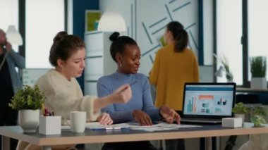 Startup employee working with african american coworker on group project comparing sales charts on clipboard with business papers on desk. Team of colleagues analzying market share looking at laptop.