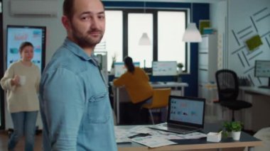 Portrait of confident man entering frame posing with arms crossed and smiling in busy startup office. Relaxed employee acting casual looking at camera in marketing department of small company.