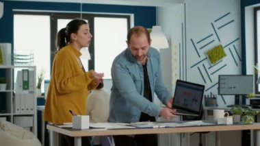 Startup office worker explaining sales startegy to new employee using laptop with business analytics and turnover charts. Man standing at desk talking about marketing with woman doing internship.