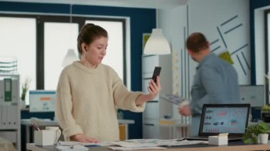 Video konferansında akıllı telefon kullanan bir kadın yeni açılan ofiste elinde çizelgelerle satış kağıtları tutmaktan bahsediyor. Uzaktan kumandalı iş arkadaşı cep telefonu kullanıyor..