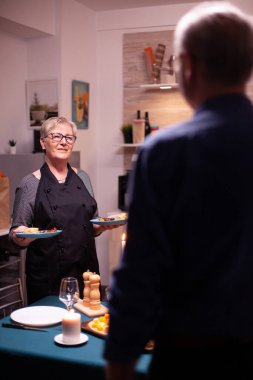 Son sınıf çifti mutfakta sohbet ediyor. Önlük giyen yaşlı bir kadın. Yaşlı çift konuşuyor, mutfakta masada oturuyor, yemeğin tadını çıkarıyor, yıldönümlerini kutluyorlar.