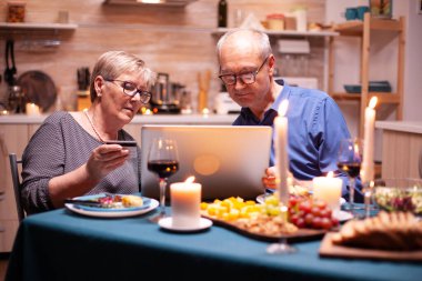 Emekli kadın romantik akşam yemeğini ödemek için kredi kartını kullanıyor. Mutfakta kocasına laptop kullanıyor. Masada oturan yaşlılar, bakıyorlar, teknolojiyi kullanıyorlar, internet kullanıyorlar..