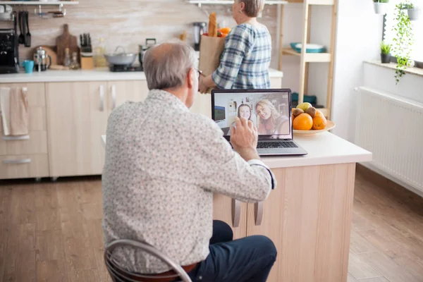 在与家人举行的视频会议上 快乐的老人在厨房里用笔记本电脑向他的侄女挥手 与女儿在线通话 使用现代网上通讯技术的长者 — 图库照片