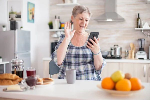 stock image Senior woman waving during video conference using smartphone in kitchen while having breaksfat. Elderly person using internet online chat technology video webcam making a video call connection camera