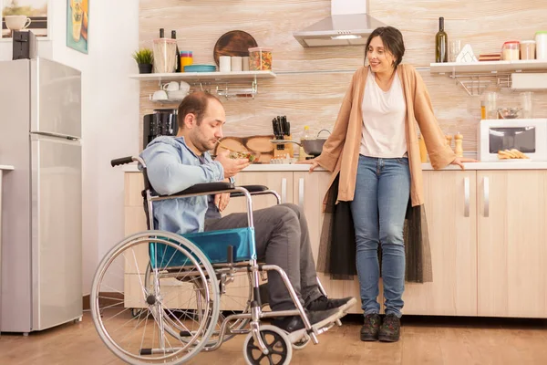 坐在轮椅上的残疾人看着妻子在厨房做的沙拉 事故后有行走残疾的瘫痪残疾人融入社会 — 图库照片