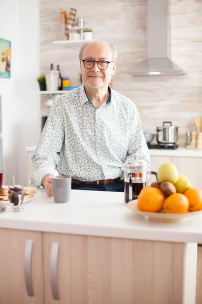 Senior man in kitchen smiling looking at camera holding hot coffee cup. Portrait of relaxed elderly older person in the morning, enjoying fresh warm drink. Healthy smiling adult face