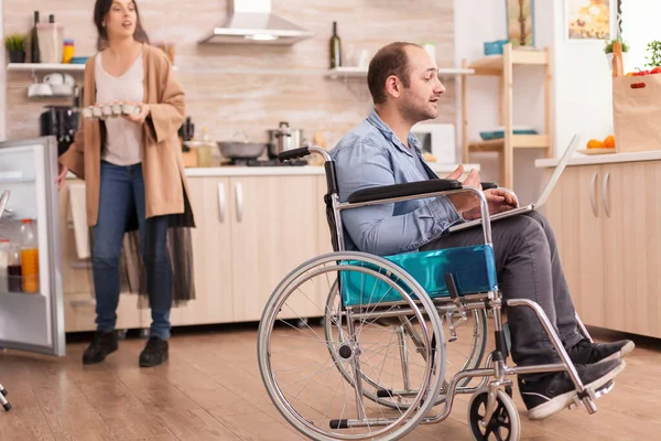 轮椅上的残疾人在厨房用笔记本电脑进行视频通话时 手里拿着一盒鸡蛋 开着冰箱门 患有行走残疾的瘫痪残疾人 — 图库照片