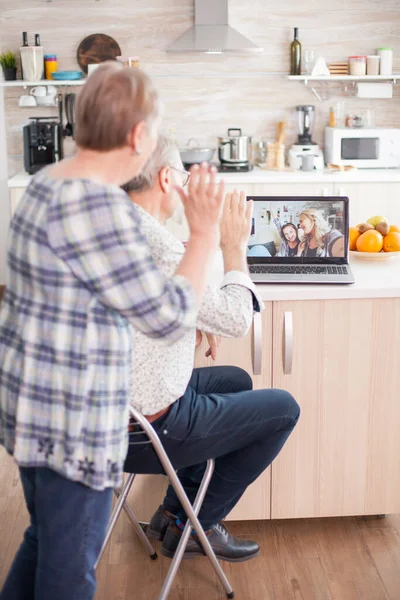 Senior couple saying hello to daughter and niece. Happy senior woman during a video conference with family using laptop in kitchen. Online call with daughter and niece. Elderly person using modern