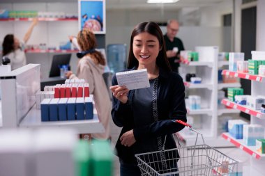 Neşeli müşteri, almayı seçmeden önce destek paketleri okuma reçetesi tutuyor. Gülümseyen müşteri eczane dükkanında sepet alışverişi yapıyor.