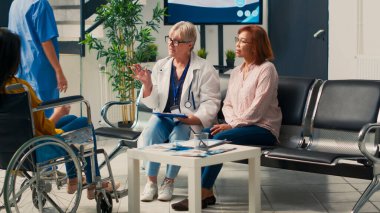 Tekerlekli sandalyedeki hasta sağlık merkezinin lobisindeki doktorla sağlık kontrolü yapıyor. Tekerlekli sandalye kullanıcısı randevusunda kronik bozukluktan muzdarip. Üçayak atışı.