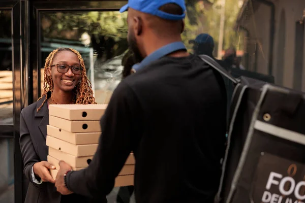 Business lunch delivery, cheerful client holding pizza boxes pile, smiling woman receiving order from courier near office building. African american employee taking meal packages stack