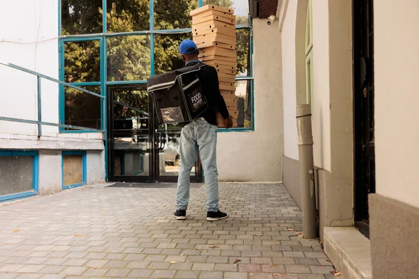 Courier carrying huge pizza boxes pile back view, deliveryman holding food order, standing near office building. Lunch delivery service african american worker delivering fastfood