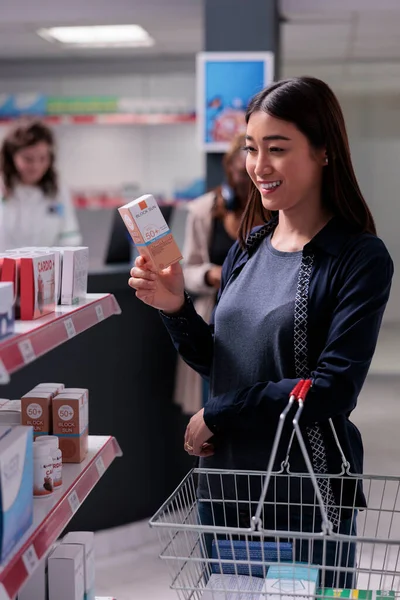 Smiling asian pharmacy customer holding basket looking at shelves with health care products buying supplements. Client choosing vitamins for immune system in pharmaceutics shop