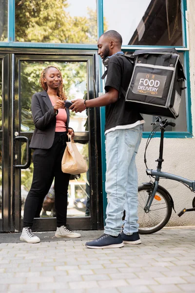 Customer paying for takeaway lunch delivery with credit card, pos terminal contactless payment for food order. African american courier with backpack delivering restaurant meal to office employee