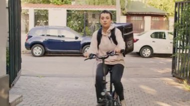 Kadın restoran çalışanı, insanların ön kapısına gidip yemek dağıtıyor, bisiklete biniyor ve müşterilere termal sırt çantasında yemek veriyor. Bisikletli taşıyıcı olarak çalışan kadın, güneşli bir gün.
