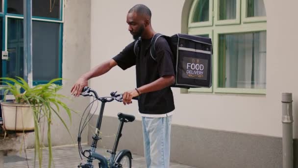 快乐的非洲裔美国信使等着外面的客户 骑自行车带着热背包送外卖 送货员站在办公大楼入口附近 看着摄像机 — 图库视频影像