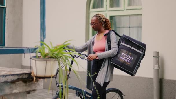 アフリカ系アメリカ人女性のキャリアのお客様に屋外では 自転車の横にある熱袋でレストランの食事の注文を提供する立って待っている バックパックを携帯し ファーストフード エクスプレスサービスを保持 — ストック動画