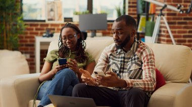Genç ortaklar koltukta birlikte mobil pohne kullanıyor, internette geziniyor ve sosyal medyada geziniyor. Aşık insanlar dijital aletlere gülüyor, boş zamanların keyfini çıkarıyor..