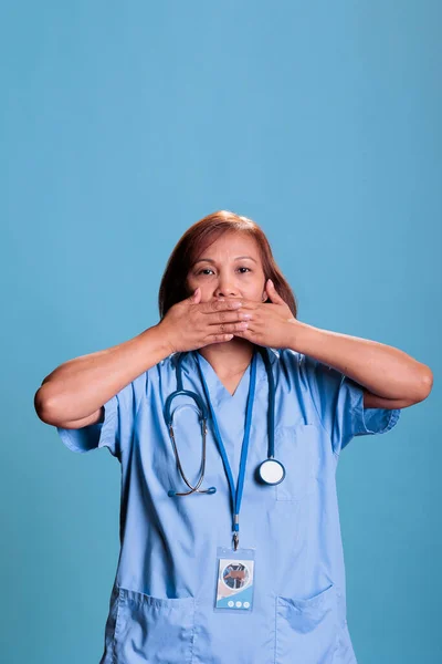 Senior nurse in medical uniform covering mouth with hands, speak no evil, three wise monkeys concept. Physician assistant working at health care expertise planning disease treatment